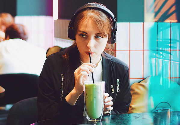 Eine Frau sitzt im Café, trinkt ein grünes Getränk über einen Strohhalm und hat Kopfhörer auf dem Kopf auf.